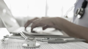 Ein Stethoskop liegt auf dem Tisch. Im Hintergrund sitzt ein medizinischer Mitarbeiter am Tisch und tippt an einem Laptop.