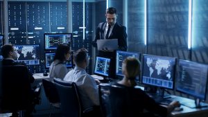 Es arbeiten fünf Menschen in einem Security Operations Center an PC Bildschirmen. Ein Mann steht mit einem aufgeklappten Laptop vor Ihnen und gibt Anweisungen.