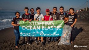 Eine Gruppe junger Menschen hält ein Plakat in der Hand "Stopping Ocean Plastic" und sammelt Müll am Strand