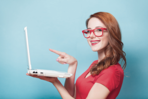 Frau mit roten Haaren und roter Brille lächelt und hält Laptop in der linken Hand während sie mit dem rechten Finger auf den Bildschirm zeigt und lächelt.